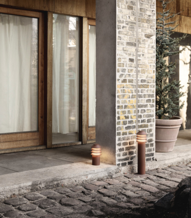 Kültéri jelenet két modern, hengeres kerti útszéli lámpával egy kő sétány és egy tégla-beton oszlop mellett. Mögöttük fából keretezett üvegajtók láthatók könnyű függönyökkel és egy nagy cserepes örökzöld növénnyel. A lámpák meleg fénye otthonos és hívogató hangulatot teremt.